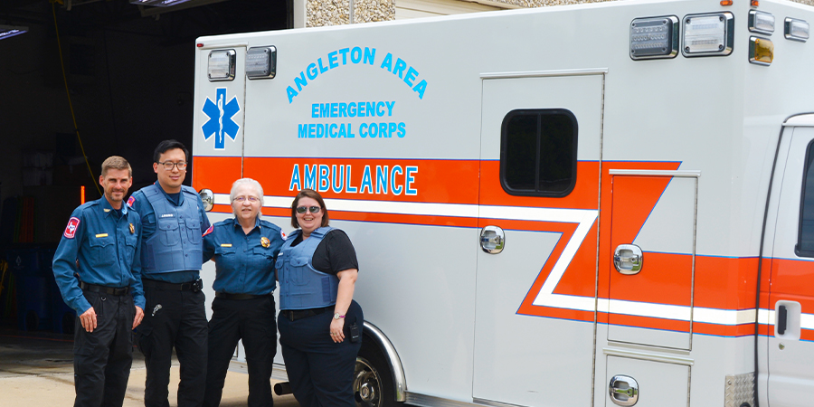 four first responders wearing uniforms with blue tops and black pants in front of an ambulance. the individuals are standing side by side