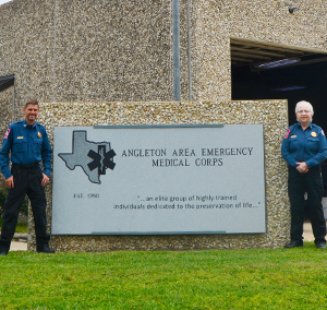 a male first responder wearing a blue shirt and black pants standing near a large concrete sign a female first responder is standing on the other end wearing a blue shirt and black pants