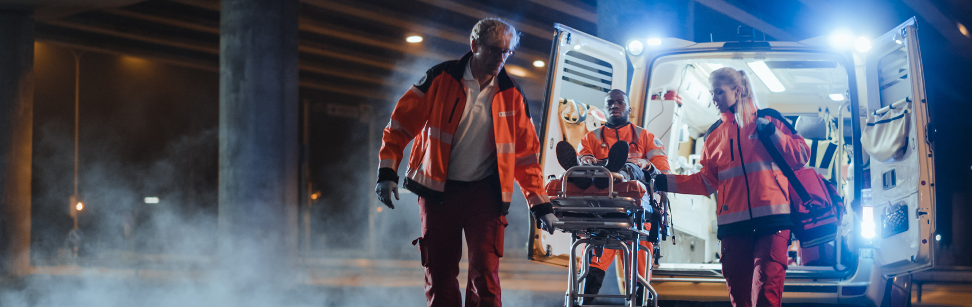 two male and one female medic wheeling a patient on a stretcher behind an opened ambulance