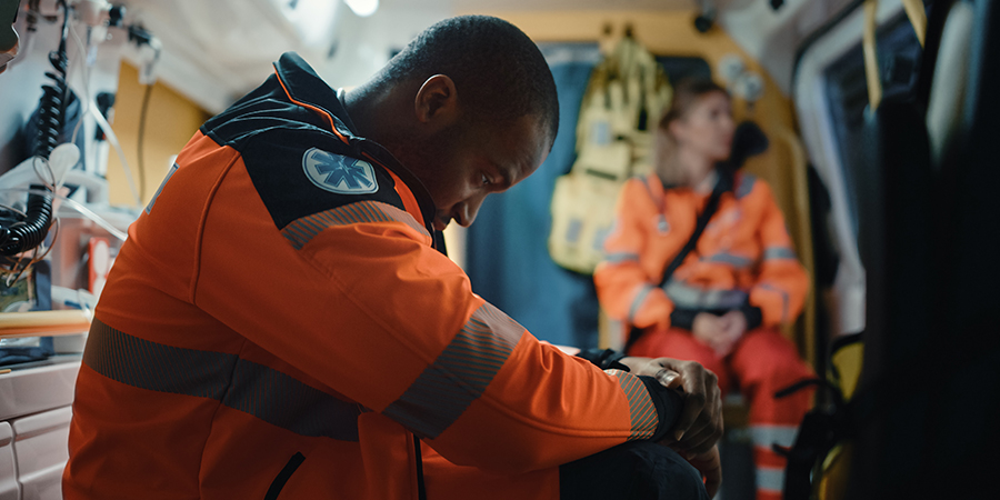 image of black male paramedic sitting in ambulance with his head hanging low. he's wearing high-visibility orange with the medic symbol on it