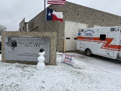 exterior of EMS building. A Texas and USA flag are flying, and there's a snowman assembled in the front. an ambulance is in the drive