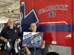 two men standing in front of a Seabrook EMS vehicle. they are holding a sign and two bags that tout "EMS Week"