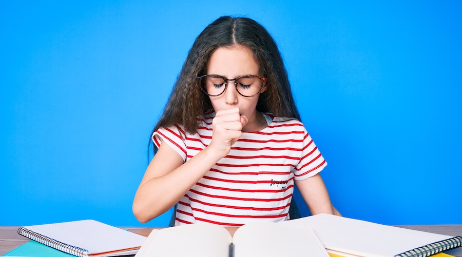 image of adolescent brunette girl with glasses and a red and white striped shirt coughing while sitting at a table covered in books and notebooks.