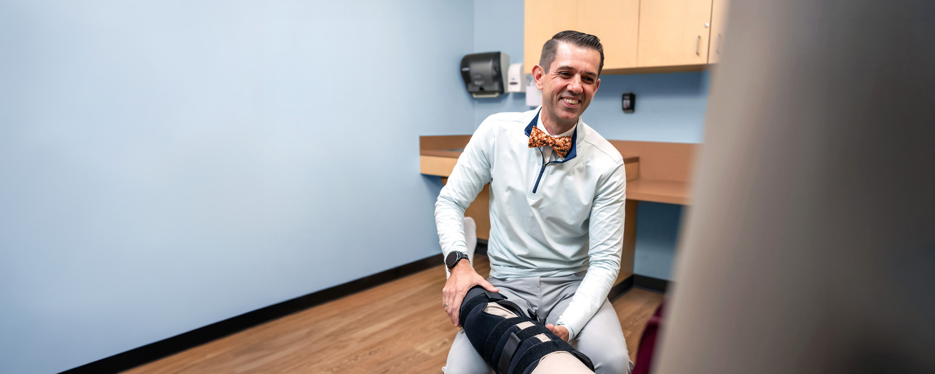 Health care provider seated in an exam room, adjusting a patient’s leg brace while the patient is partially visible in the foreground.