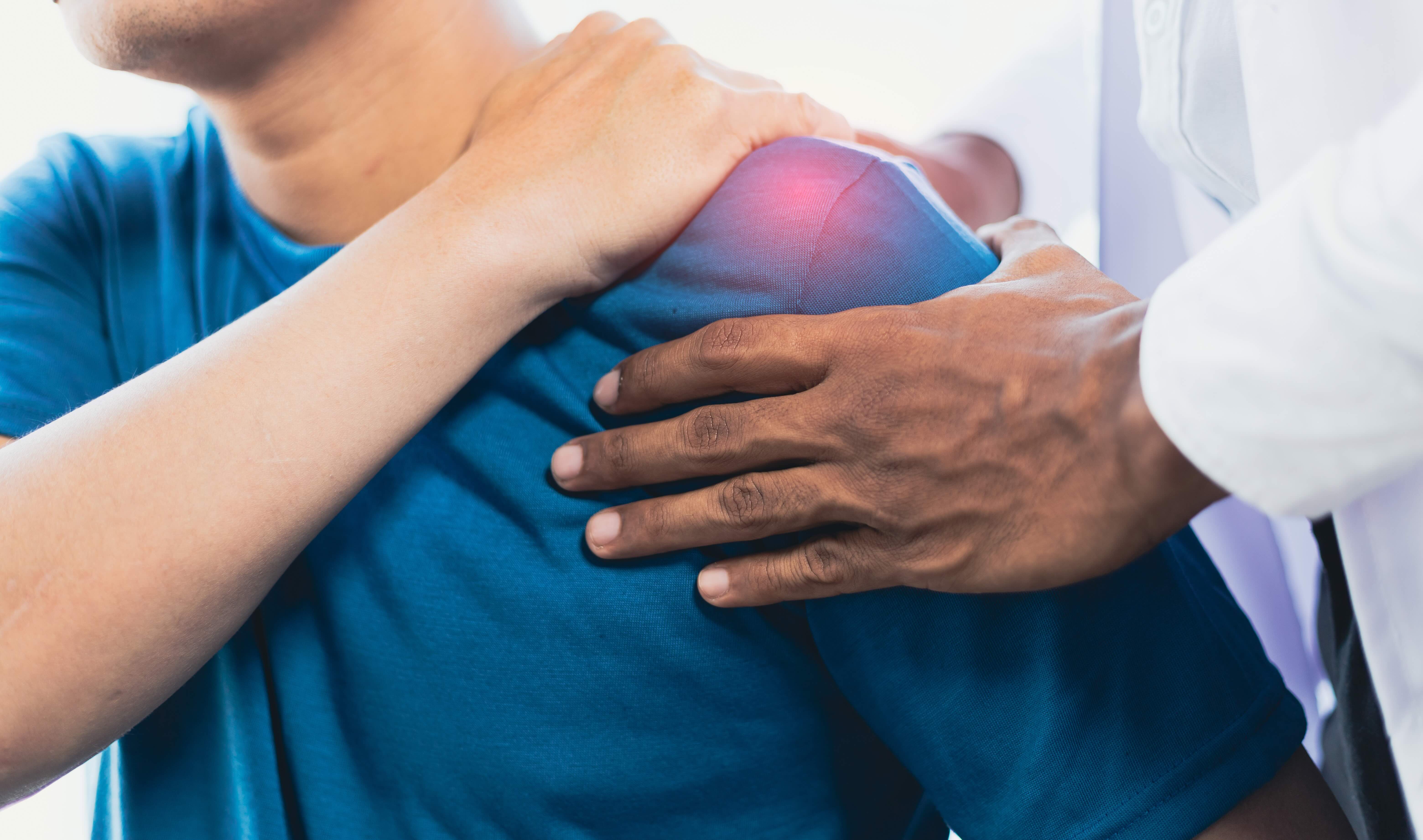 Close-up of a health care provider examining a person’s shoulder, with one hand placed on the upper arm and shoulder area highlighted to indicate pain or injury during a medical evaluation
