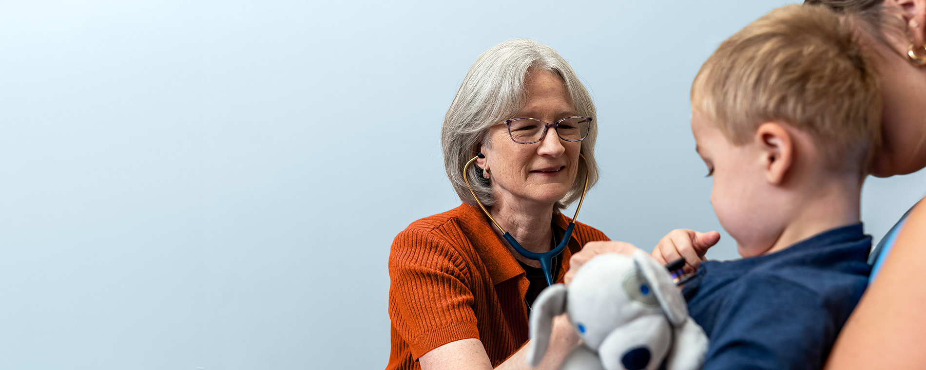 Clinician uses a stethoscope to examine a young child while a caregiver holds the child, with a stuffed toy visible in the foreground.Provide your feedback on BizChat