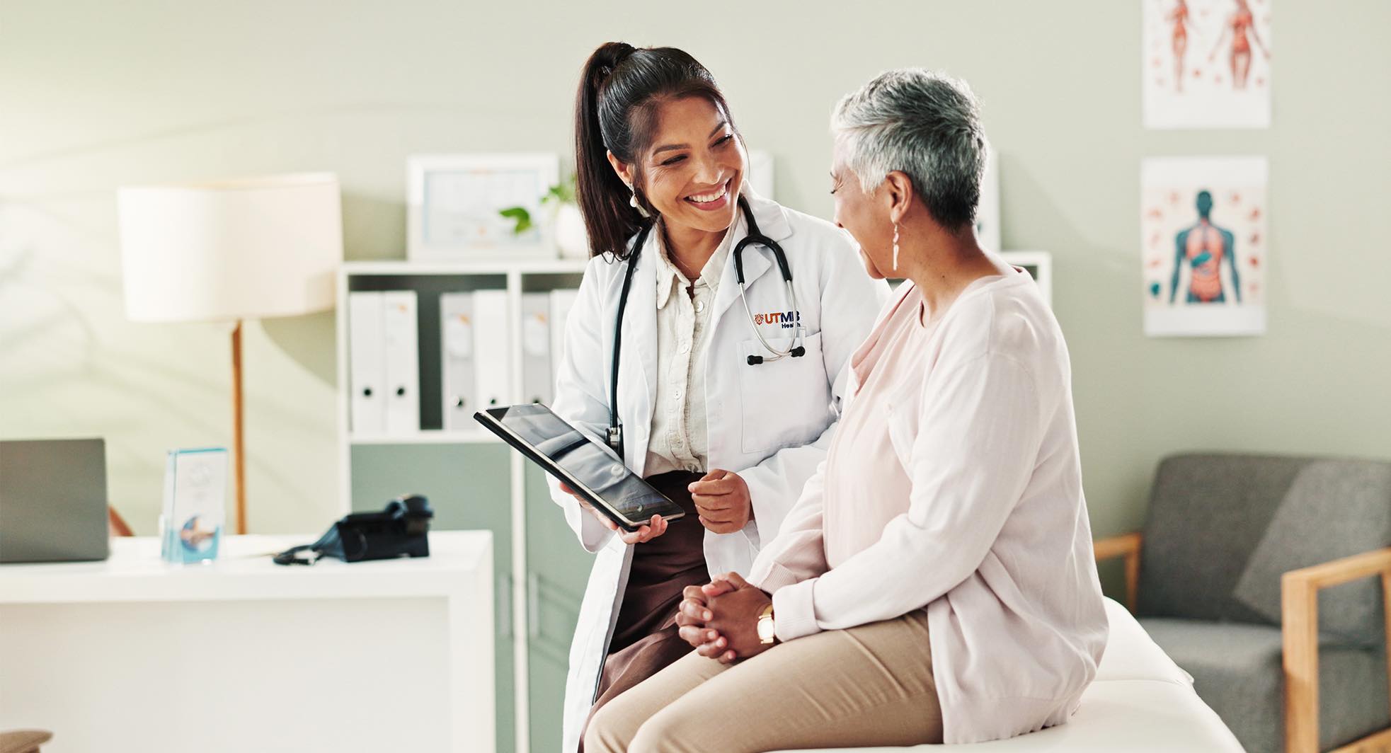 Female doctor with brown hair and tablet smiling while talking to an older female patient.