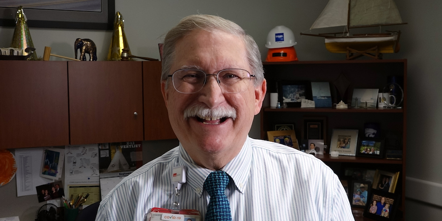 Person wearing glasses and a tie seated in an office with shelves, framed photos and office items in the background