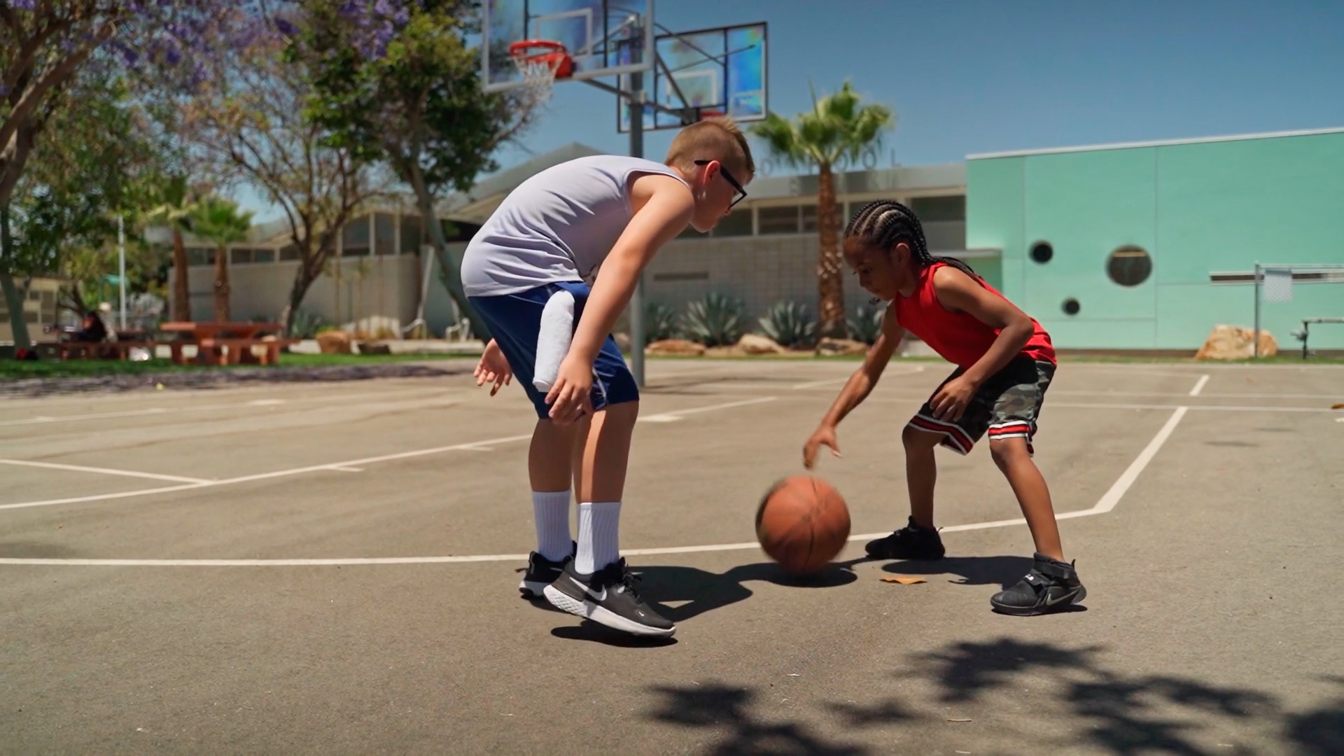 Two children play basketball on an outdoor court under bright sunlight, with a hoop and school building in the background, illustrating physical activity during warm weather
