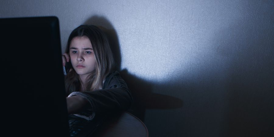 forlorn adolescent girl on phone in dark room behind laptop screen