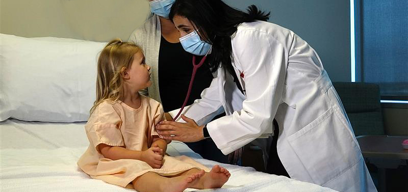 A child sits on a hospital bed while a doctor wearing a white coat uses a stethoscope to listen to the child’s heart, with another adult standing nearby in a patient room.