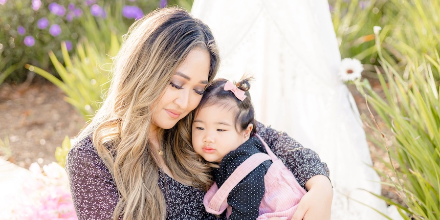 an asian mom holding her young daughter. they are leaning in toward each other and lovely green plants are in the background