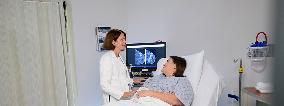 Clinician in a white lab coat speaks with a patient lying in a hospital bed, with medical imaging displayed on a monitor nearby inside a clinical exam room