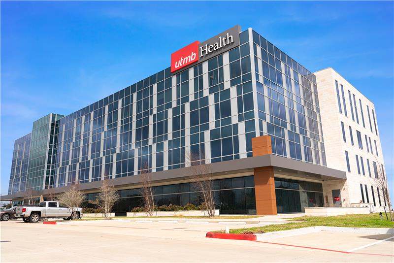 Exterior view of UTMB Health’s Primary and Specialty Care Clinic in Friendswood, showing a modern multi‑story building with a glass façade, the UTMB Health logo, parking area and landscaped surroundings under a clear sky