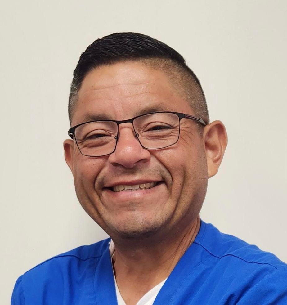 close-up headshot image of Juan Tovar wearing blue scrub shirt with white undershirt. He has black, short-cut hair and subtle black-frame glasses