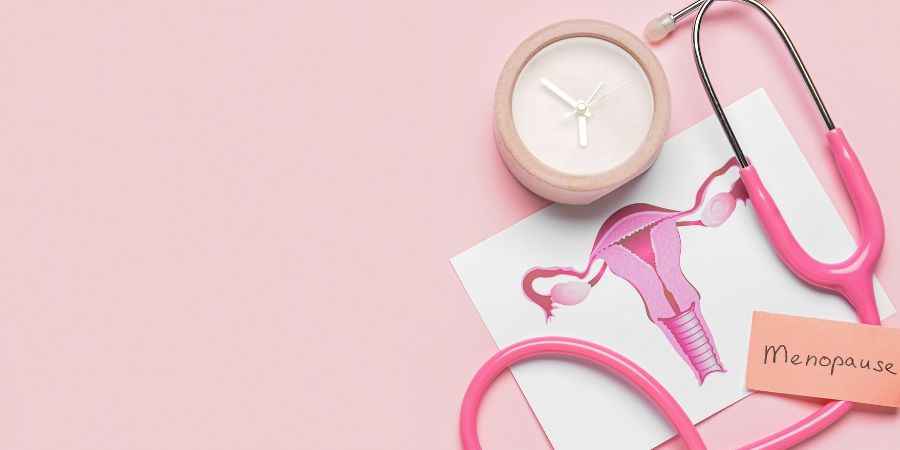 Pink background with a stethoscope, an illustration of the female reproductive system, a small clock, and a note labeled “Menopause,” symbolizing hormonal changes over time