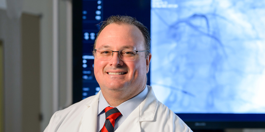 Person wearing a white lab coat, glasses and a striped tie standing in a clinical setting, with medical imaging displayed on monitors in the background