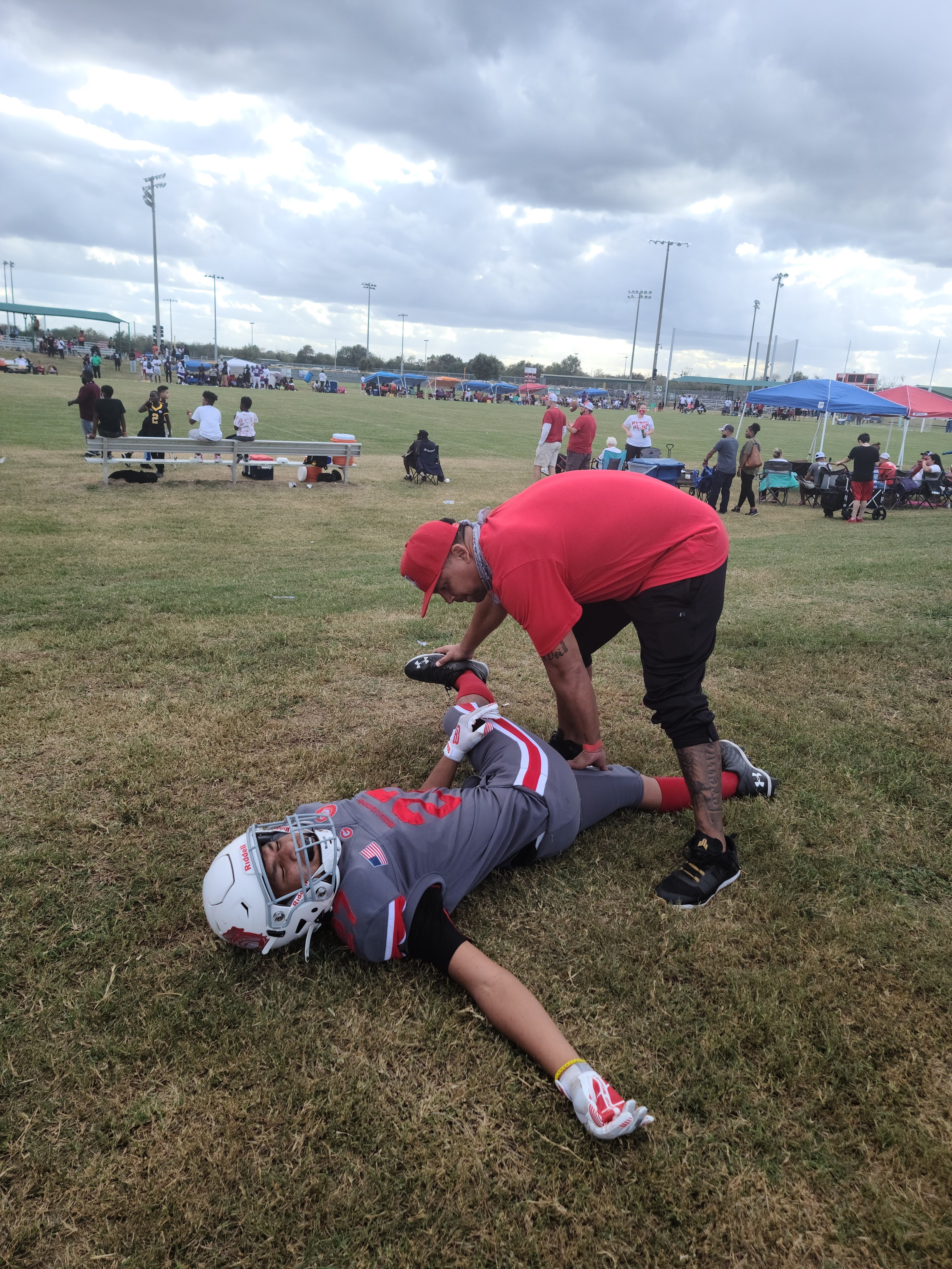 image of UTMB Health patient Priest Simpson stretching on the sidelines of a football field in his football uniform with a coach assisting him with his leg