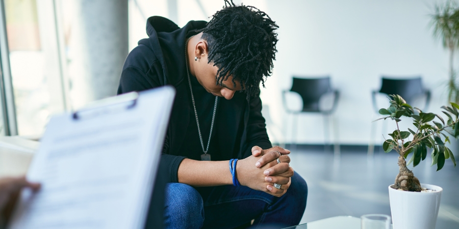 a clipboard with a white piece of paper is in the foreground, blurry. a teen in a black hoody and jeans sits with his head hunched over and hands clasped, resting on his legs. a plant in a white pot is to the right. there's 2 blurry chairs in the BG