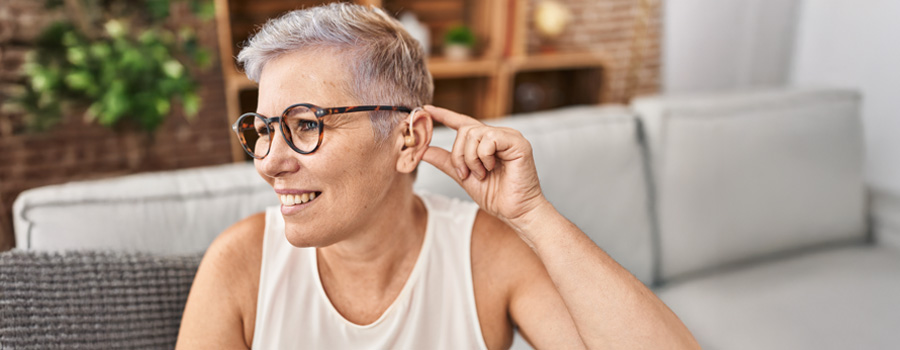 A smiling woman adjusts her behind-the-ear hearing aid while sitting at home.