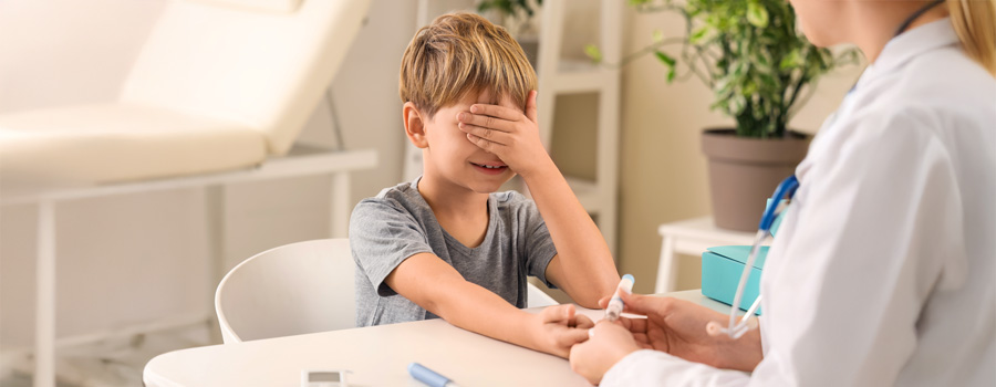 Little diabetic boy covering his eyes while doctor uses lancet pen to check his glucose at table in clinic