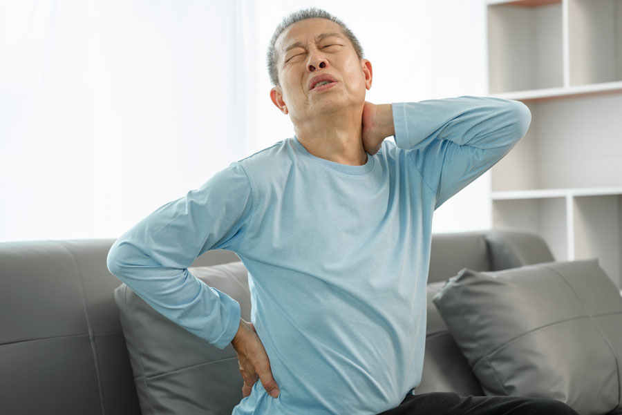 Elderly man sitting on sofa, grimacing from lower back pain and neck pain as he touches his neck and back
