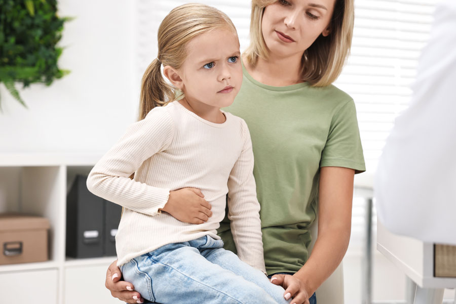 Young girl with troubled expression touching her belly while seated on her mother’s lap at hospital