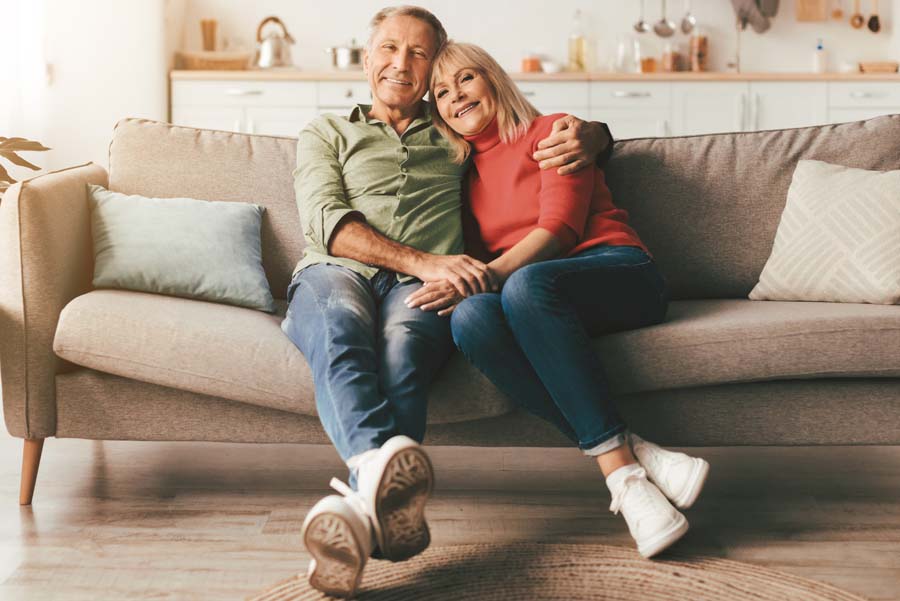 Happy Mature Couple Embracing Posing Sitting On Couch At Home, Smiling Looking At Camera. Retirement And Family Lifestyle, Marriage And Happiness Concept