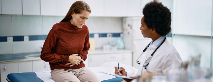 Woman on hospital bed touching her belly, experiencing stomach pain, as female doctor takes notes on clipboard