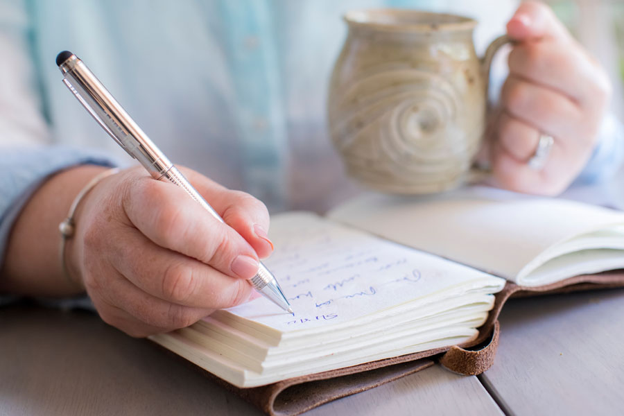 A person writes in a notebook while holding a mug highlighting fine motor skills and daily function affected by movement disorders.