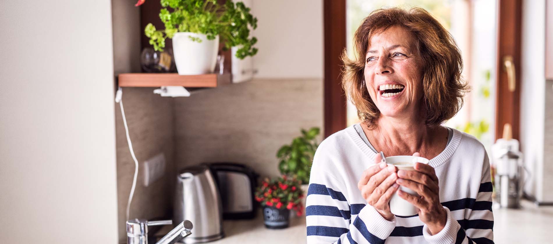 A woman smiles at home while holding a warm drink representing daily life and comfort while living with a movement disorder.