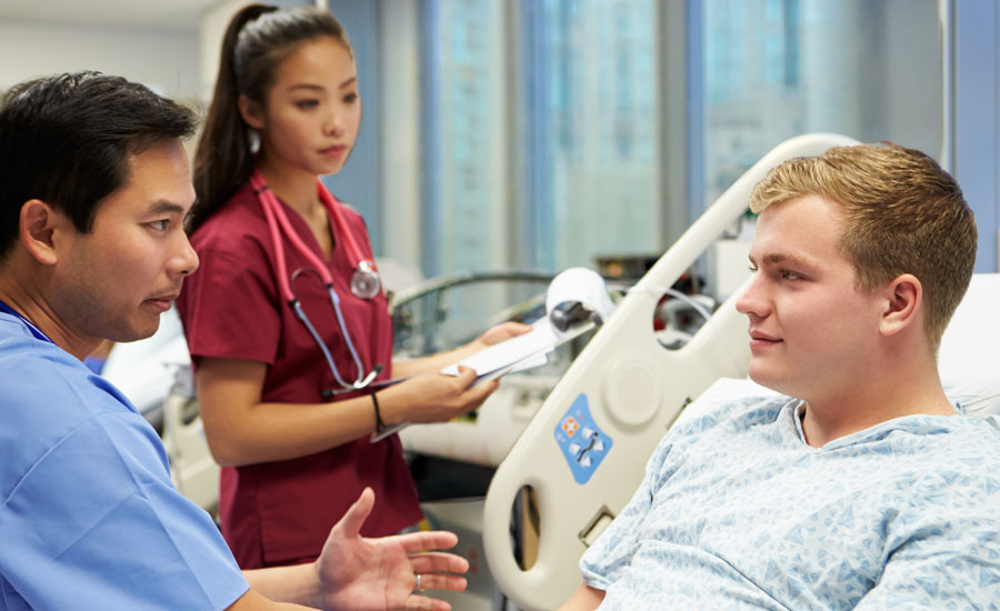 Male doctor speaking with young man lying on bed in hospital emergency room