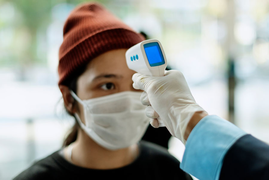 Young woman wearing a hat and mask, getting her temperature checked with an electronic thermometer in medical setting