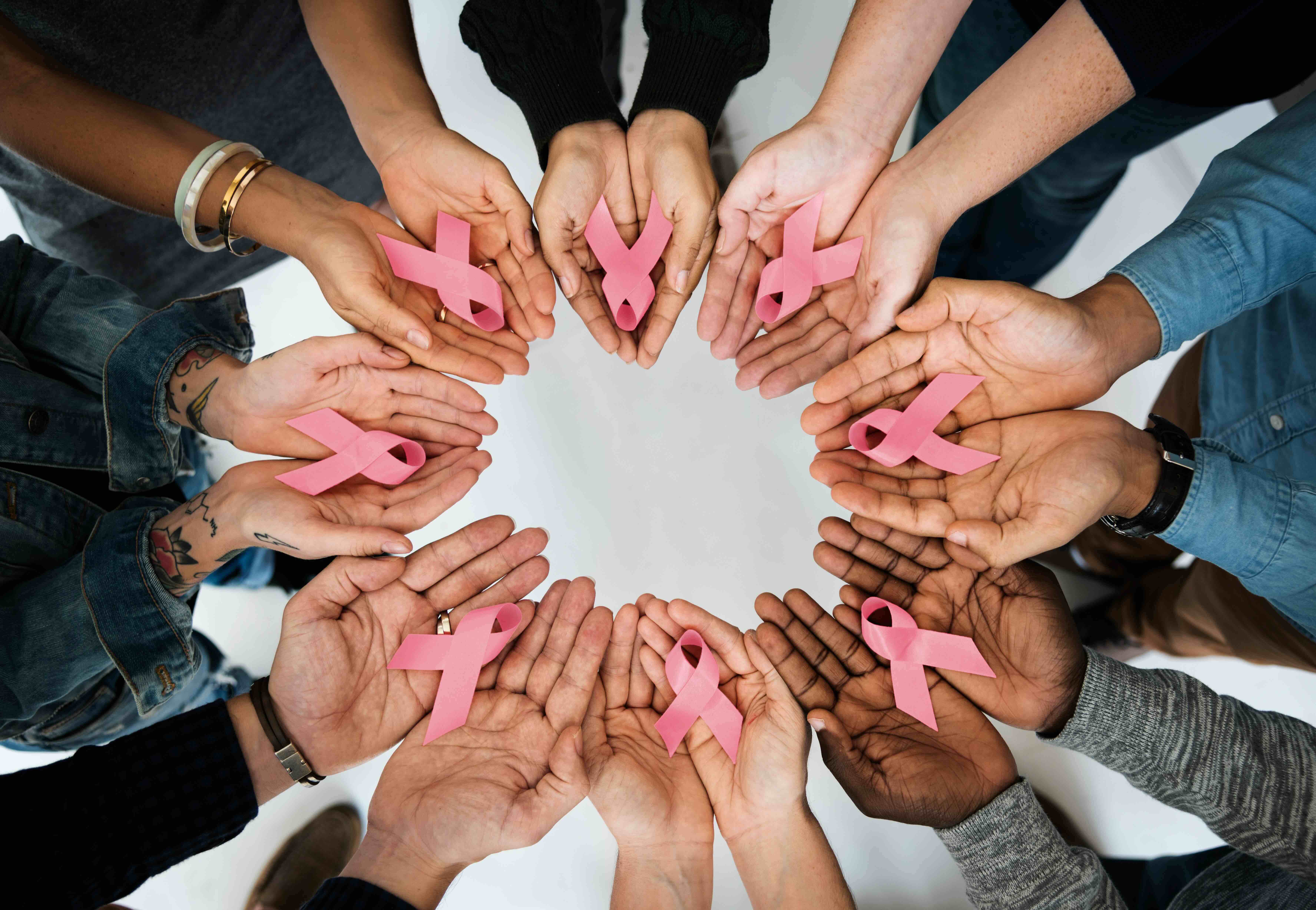 Multiple hands arranged in a circle, each holding a pink breast cancer awareness ribbon against a light background