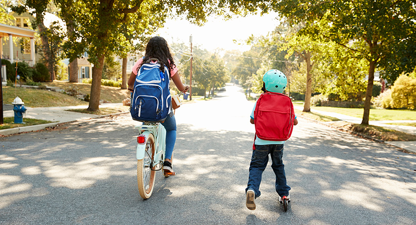 Two children with backpacks travel down a tree-lined residential street, one riding a bicycle and the other on a scooter while wearing a helmet.