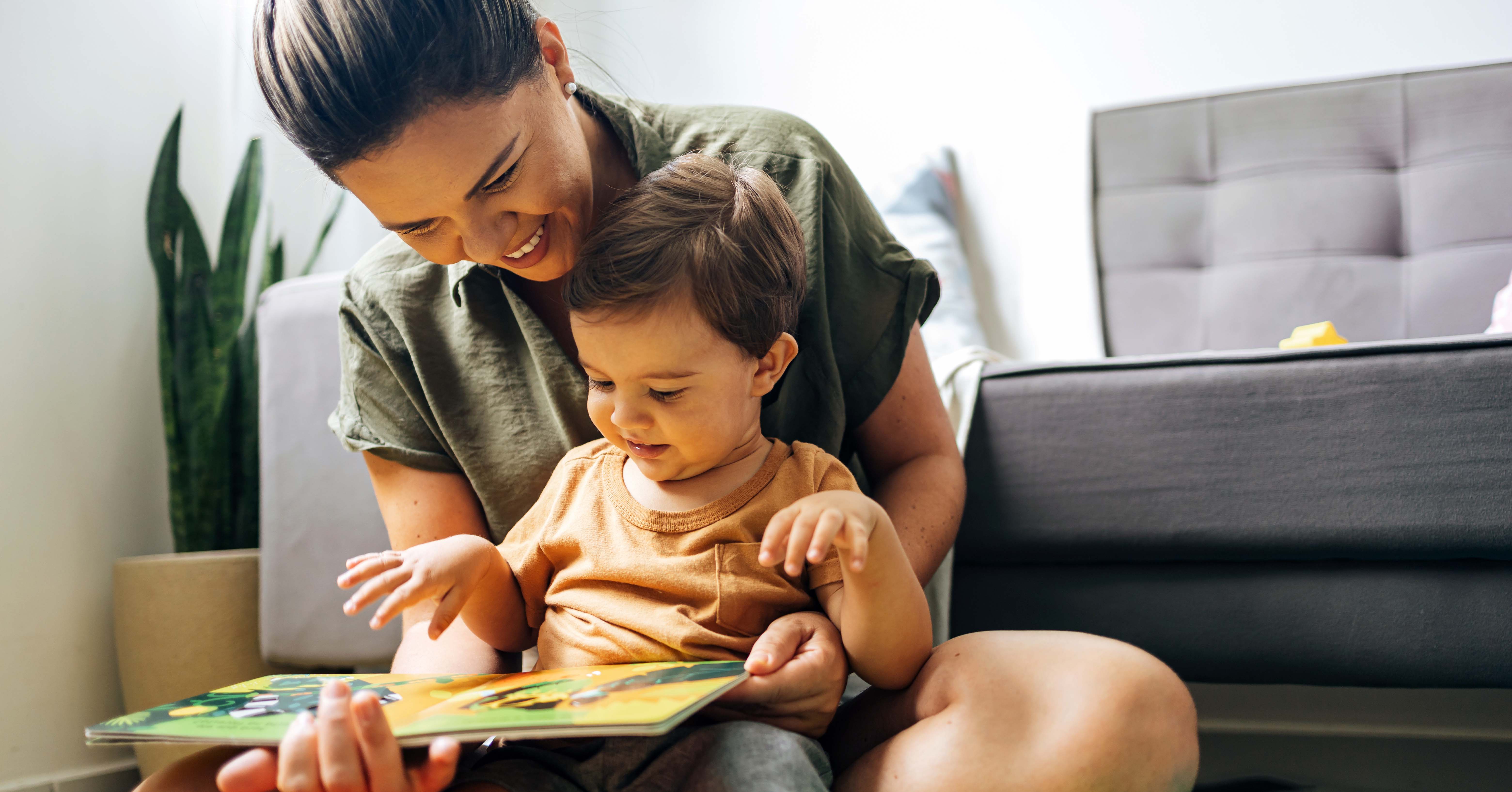 An adult and a toddler sit together on the floor of a living room, looking at an open picture book, with a couch and houseplant visible in the background