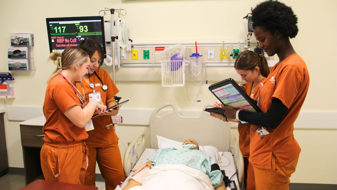 Nursing students in orange scrubs use tablet devices while practicing clinical skills around a hospital bed with a medical mannequin in a simulation lab.