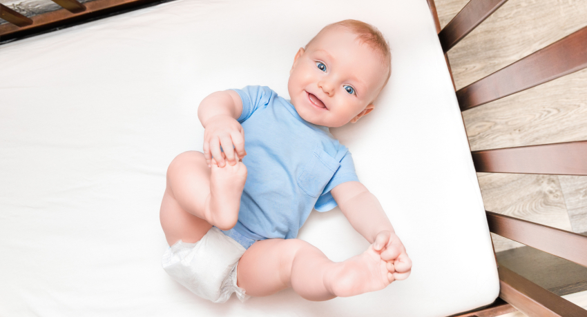 Smiling baby lying on a white mattress, wearing a blue shirt and diaper, with arms and legs raised.