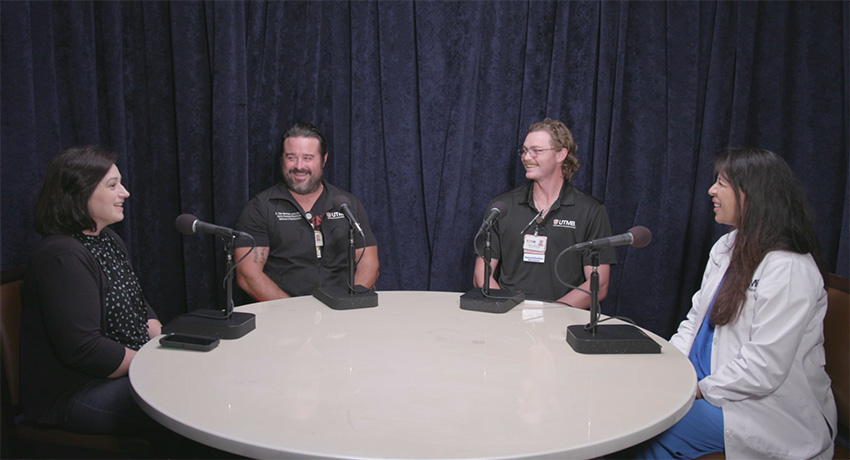 Four people sit around a round table with microphones, participating in a recorded discussion in front of a dark curtain backdrop. Two participants wear UTMB-branded shirts, and one participant wears a white coat.