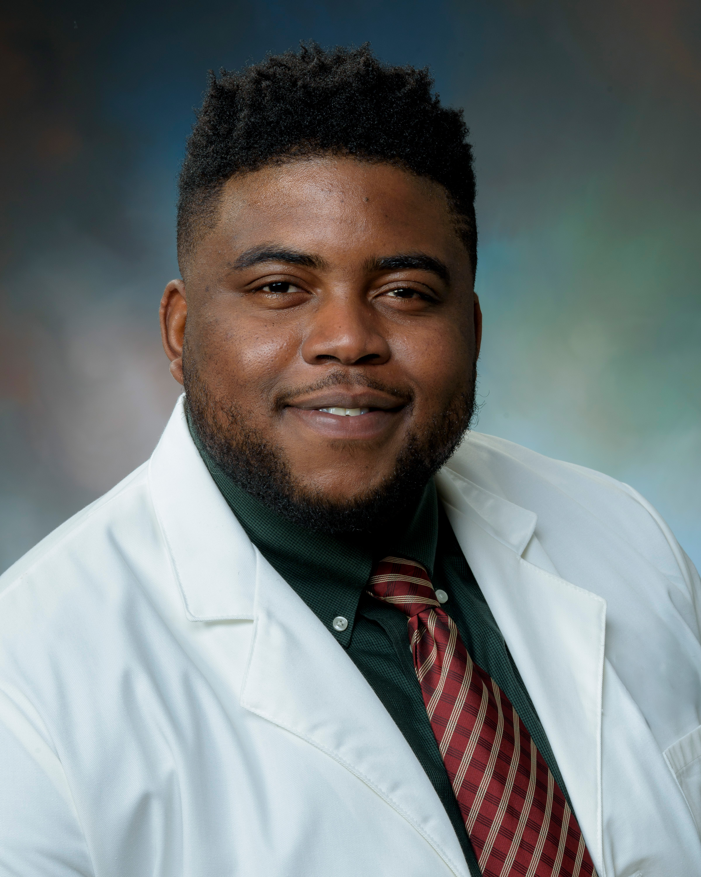 Studio headshot of an Dr. Ben-Okoli wearing a white lab coat, collared shirt, and patterned tie, looking at the camera against a blue‑green gradient background.