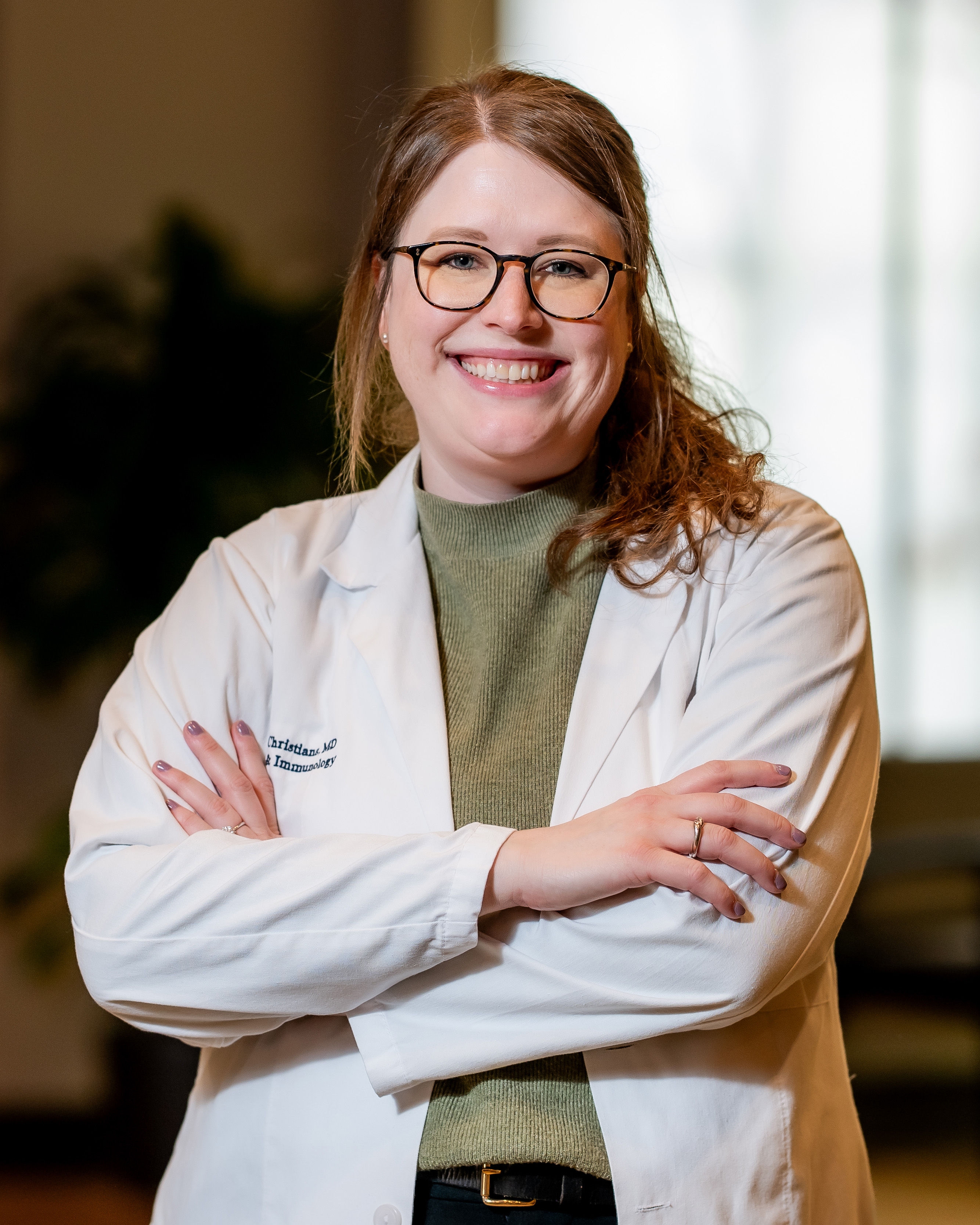 Dr. Christians Healthcare professional in a white coat with a UTMB Health logo, wearing glasses and standing with arms crossed indoors against a softly blurred background.