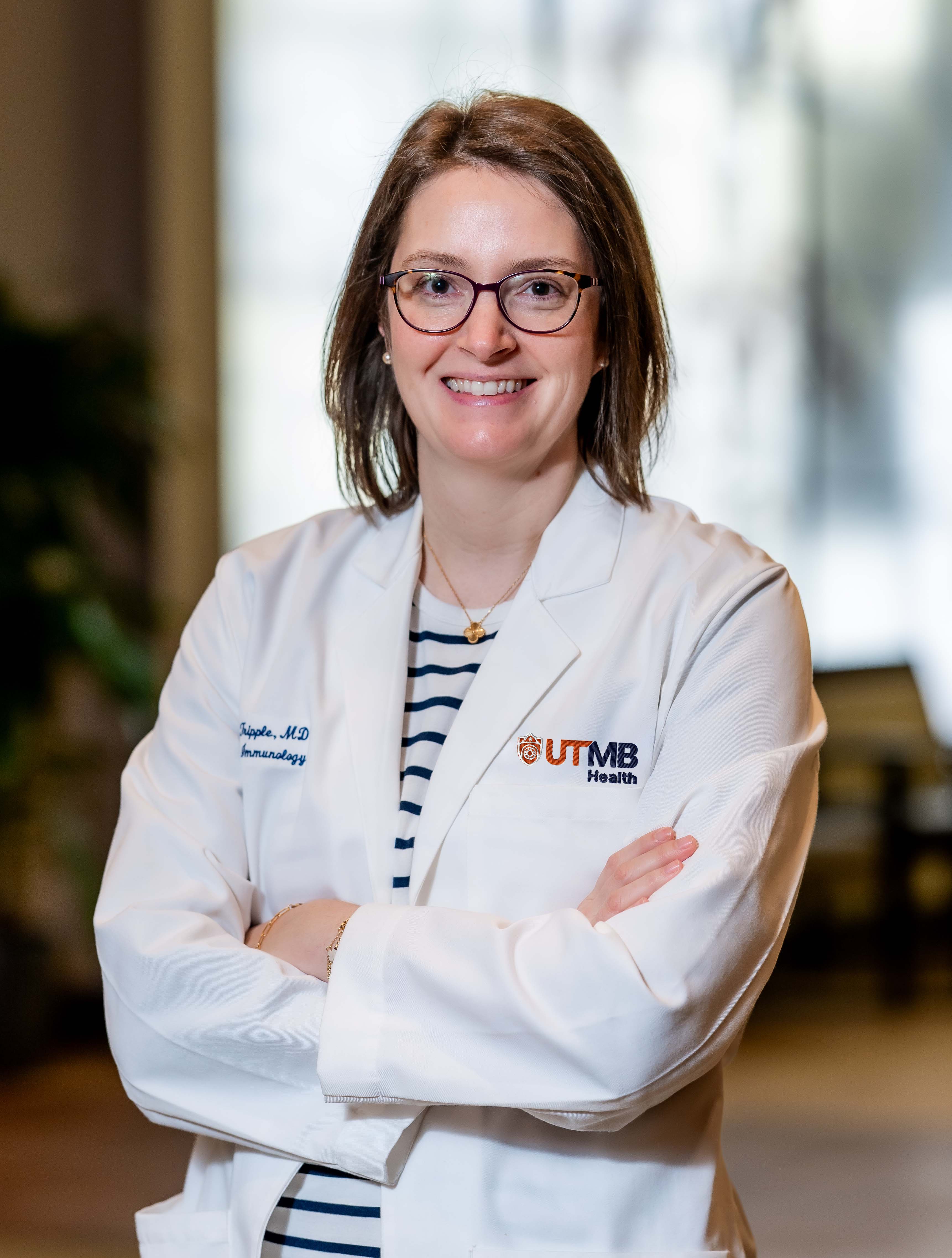 A person wearing a UTMB Health white lab coat stands indoors with arms crossed, shown from the waist up against a softly blurred background.