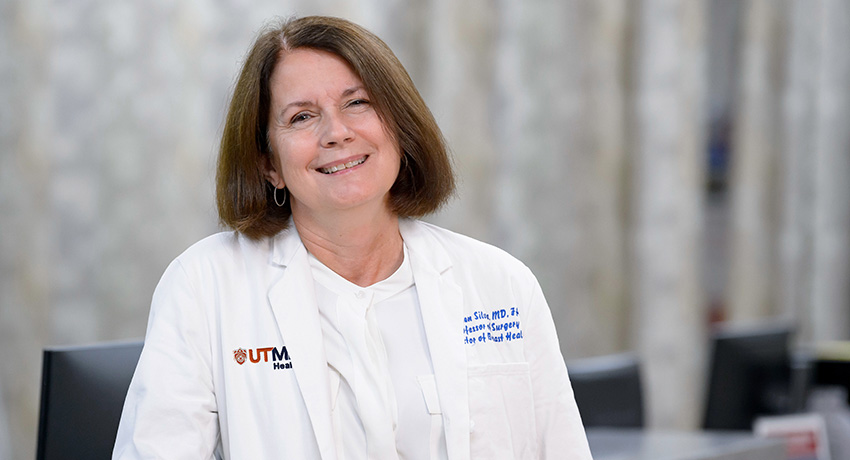 Person wearing a white UTMB Health lab coat sits indoors facing the camera. The coat displays a UTMB Health logo and embroidered professional credentials.