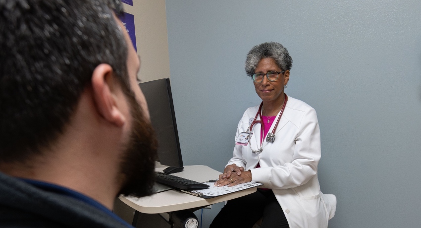 A physician wearing a white coat and stethoscope sits across from a patient during an appointment in a medical exam room. The doctor appears engaged and attentive while speaking with the patient beside a computer workstation.