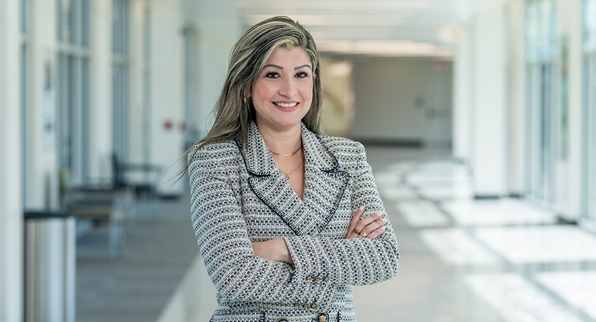 Person wearing a patterned jacket stands with arms crossed in a bright hallway with glass walls.