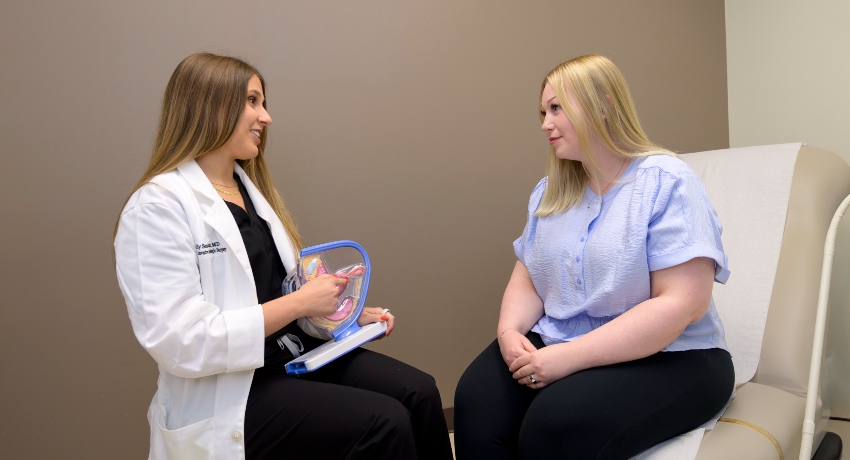 A healthcare provider in a white coat sits facing a patient in an exam room, using a plastic anatomical model to explain a medical concept. The patient, seated on an exam table, listens attentively as they engage in a calm, one-on-one discussion.