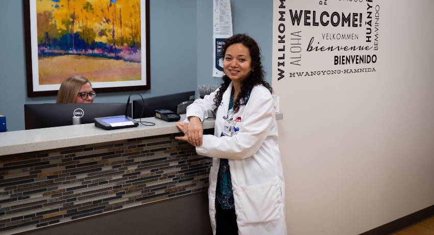 A doctor in a white coat stands at a clinic reception desk, smiling toward the camera while leaning on the counter. A receptionist works behind the desk, and a wall display featuring the word “Welcome” in multiple languages is visible in the background.