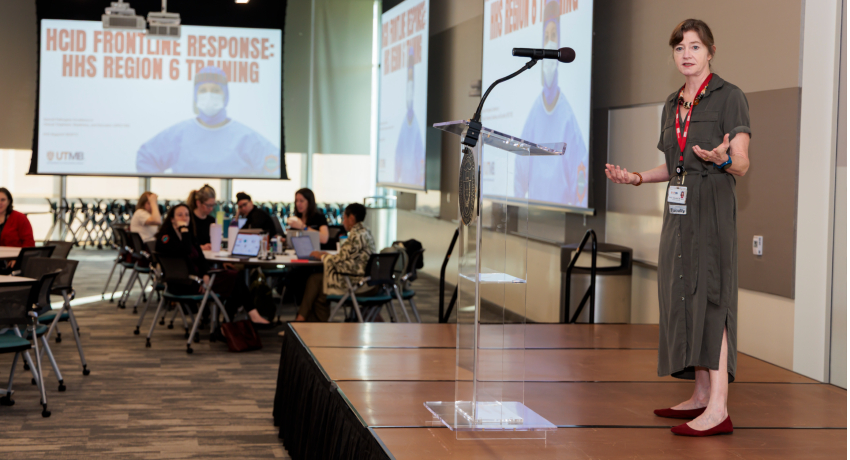A woman wearing a brown dress and a UTMB lanyard stands at a clear podium addressing seated attendees at round tables, with a projected slide reading “HCID Frontline Response: HHS Region 6 Training” behind them.