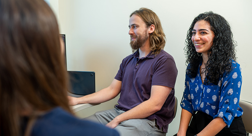 Two people sit side by side in an office or exam room, facing a third person in the foreground. A computer monitor is visible beside them.