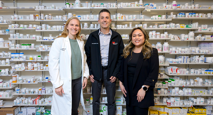 Three pharmacy team members standing in front of shelves filled with prescription medication bottles.